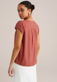 Blusa corta color ruggine con un sottile motivo testurizzato, scollo rotondo e una leggera caduta sul retro, indossata con una gonna bianca.
