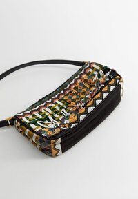 Mała prostokątna torebka na ramię z czarnym dnem, wielokolorowymi koralikami w kształcie geometrycznym, frędzlami i wąskim czarnym paskiem na tle białej powierzchni.