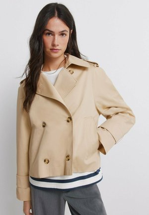 Femme aux longs cheveux noirs portant une veste beige croisée courte, une chemise blanche à rayures noires et un pantalon gris sur fond blanc.