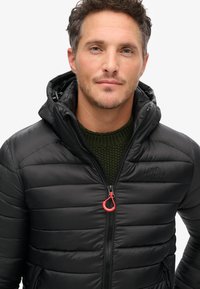 Giacca a vento nera con cappuccio, caratterizzata da una texture trapuntata, una zip frontale con tirante rosso e uno strato sottostante in maglia testurizzata verde.