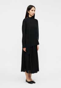 Μαύρο φόρεμα με μακριά μανίκια, ψηλό γιακά και πλισέ φούστα· διάφανο ύφασμα, εφαρμοστό μπούστο και παπούτσια μπαλαρίνες που ταιριάζουν ολοκληρώνουν την εμφάνιση.