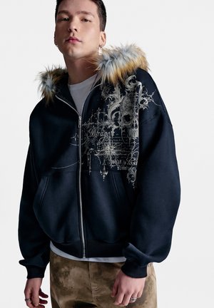 Junger Mann trägt eine schwarze Jacke mit Reißverschluss und pelzbesetzter Kapuze mit einem aufwendigen Totenkopf- und Schrift-Design vorne, beige Hose und Ringe an den Fingern.