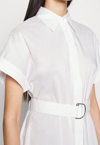 Abito bianco in tessuto morbido con maniche corte e colletto a punta. Presenta una vita con cintura e una fibbia argento per dare struttura.