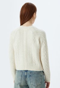 Cremefarbener Zopfmusterpullover mit ribbeligem Saum und Bündchen. Hat einen runden Ausschnitt und ein strukturiertes Muster, hergestellt aus weichem Garn.