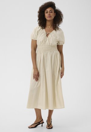 Woman with curly hair wearing a beige midi dress with puff sleeves, smocked waist, and black heeled sandals, standing against a plain background.