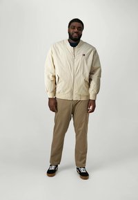 Lys beige bomberjakke med ribbet mansjetter, glidelås foran og lite logo. Kombinert med khakibukser og svarte sneakers med hvite detaljer.