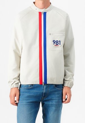 Personne portant une veste légère avec des rayures verticales rouges et bleues le long de la fermeture éclair et un patch sur la poche poitrine intitulé "981 Replay."