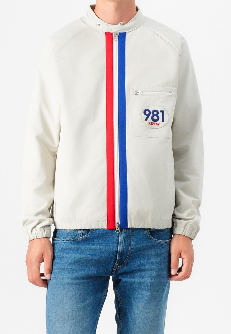 Personne portant une veste légère avec des rayures verticales rouges et bleues le long de la fermeture éclair et un patch sur la poche poitrine intitulé "981 Replay."
