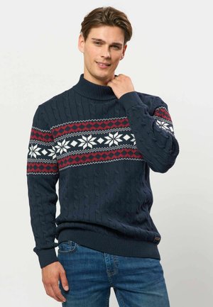 Junger Mann trägt einen dunkelblauen festlichen Pullover mit weißen Schneeflocken und roten Mustern, steht mit einer Hand nahe am Hals und lächelt.