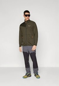 Olive rohelise lukuga jakk mustade aksentidega, mille juurde sobivad hallid püksid mustade paneelidega. Modell kannab päikeseprille ja tekstuuriga jalatseid.