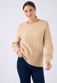 Beige kabelgebreide trui met een ronde halslijn, ribgebreide mouwen en zoom. Heeft een textuurdesign en een ontspannen pasvorm. Gecombineerd met donkere jeans.
