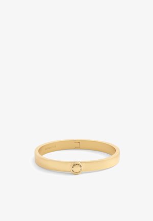 Pulsera de metal en tono dorado con un diseño plano y suave, y un cierre circular con el nombre de la marca grabado, que ofrece un aspecto minimalista.
