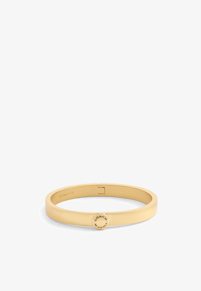 Bracelet en métal doré avec un design lisse et plat, et un fermoir circulaire portant le nom de la marque gravé, offrant un look minimaliste.