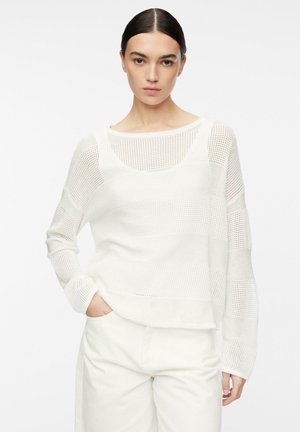 Frau mit dunklem Haar in einem weißen Netz-Langarmpullover und weißen Hosen, stehend mit einer Hand in der Tasche vor einem schlichten Hintergrund.