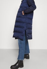 Marineblå pufferjakke med vandret syninger, lynlåslukning, sidelommer og ribbede manchetter. Matchet med lyseblå jeans og sorte ankelstøvler.