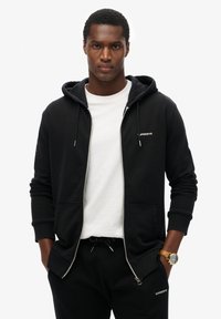 Felpa nera con zip e cappuccio, con lacci, tasche frontali e logo "Superdry". Indossata sopra una t-shirt bianca e abbinata a pantaloni sportivi neri.