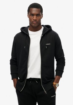 Felpa nera con zip e cappuccio, con lacci, tasche frontali e logo "Superdry". Indossata sopra una t-shirt bianca e abbinata a pantaloni sportivi neri.