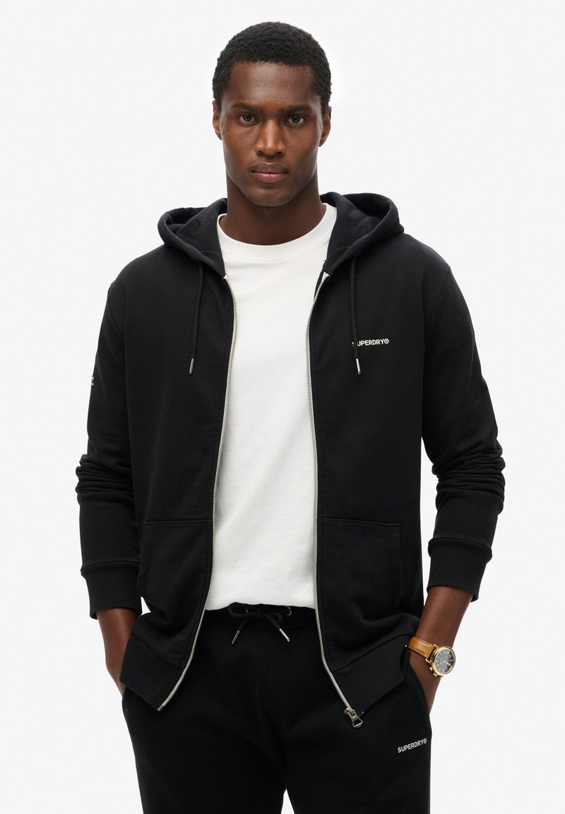 Sudadera negra con cremallera, cordones, bolsillos frontales y logo de "Superdry". Lleva una camiseta blanca debajo y combina con pantalones de chándal negros.