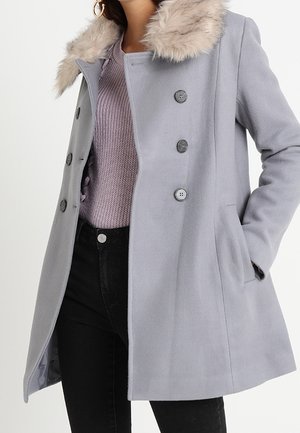 Light gray double-breasted coat with fur collar, worn over a lavender knit sweater and black pants, model’s hand in coat pocket.