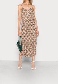 Woman wearing a sleeveless, patterned midi dress with a pink and green floral design, holding a dark green handbag, and wearing white sandals.