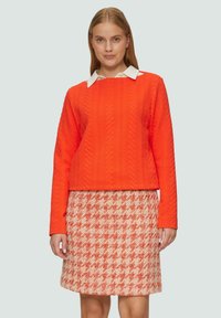 Pull en tissu texturé orange avec un col blanc et une jupe à motif pied-de-poule rose. Design simple et structuré avec des manches longues.