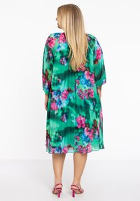 Floral midi jurk met een levendige groene basis en roze en blauwe bloemen, korte mouwen en een vloeiende, geplooide zoom. G gedragen met roze haksandalen.