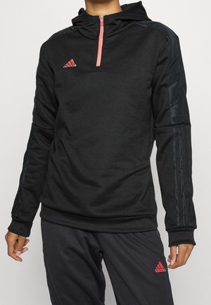 Persoon gekleed in een zwart Adidas-hoodie met rode ritssluiting en logo, gecombineerd met bijpassende zwarte Adidas-broek met rood logo.
