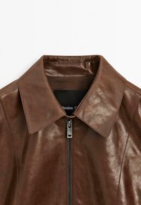 Veste en cuir marron avec un col pointu, fermeture éclair à l'avant et finition texturée. Étiquette intérieure visible avec le nom de la marque.