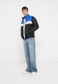 Isoleret pufferjakke med farveskift-design i sort, blå og hvid, lynlåslukning og høj krave. Bæres med lyse denimjeans.