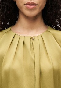 Blouse vert olive avec des plis au niveau de l'encolure et une petite fermeture boutonnée au centre. Tissu doux et lisse avec une coupe structurée.