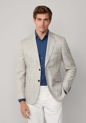 Mann trägt ein hellbeiges karierter Blazer, ein blaues Hemd mit Knöpfen und weiße Hosen. Er steht mit einer Hand in der Tasche vor einem schlichten Hintergrund.