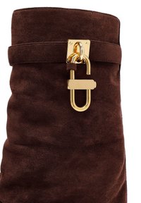 Brown suede handbag featuring a gold metal fastener with a circular design. The texture is soft, and the overall shape is rounded and structured.