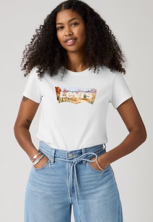 Femme aux cheveux bouclés portant un t-shirt blanc Levi's avec un logo paysage désertique et un jean taille haute bleu clair avec ceinture nouée, mains dans les poches.