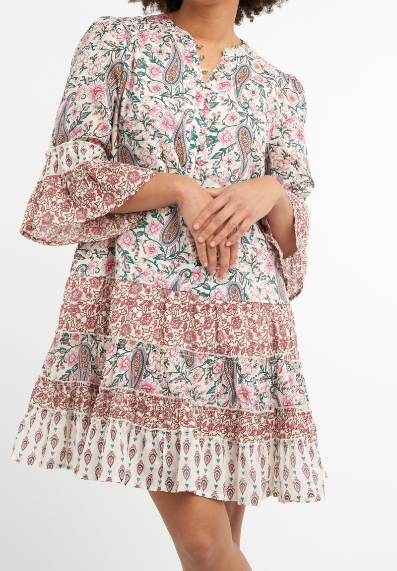 Femme portant une robe à motifs floraux et cachemire avec des manches cloche volantées et une jupe à étages, mains doucement jointes devant.