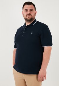 Námornícky polo tričko s krátkymi rukávmi, bielym lemovaním na golieri a rukávoch, a malým vyšívaným logom na prsiach.