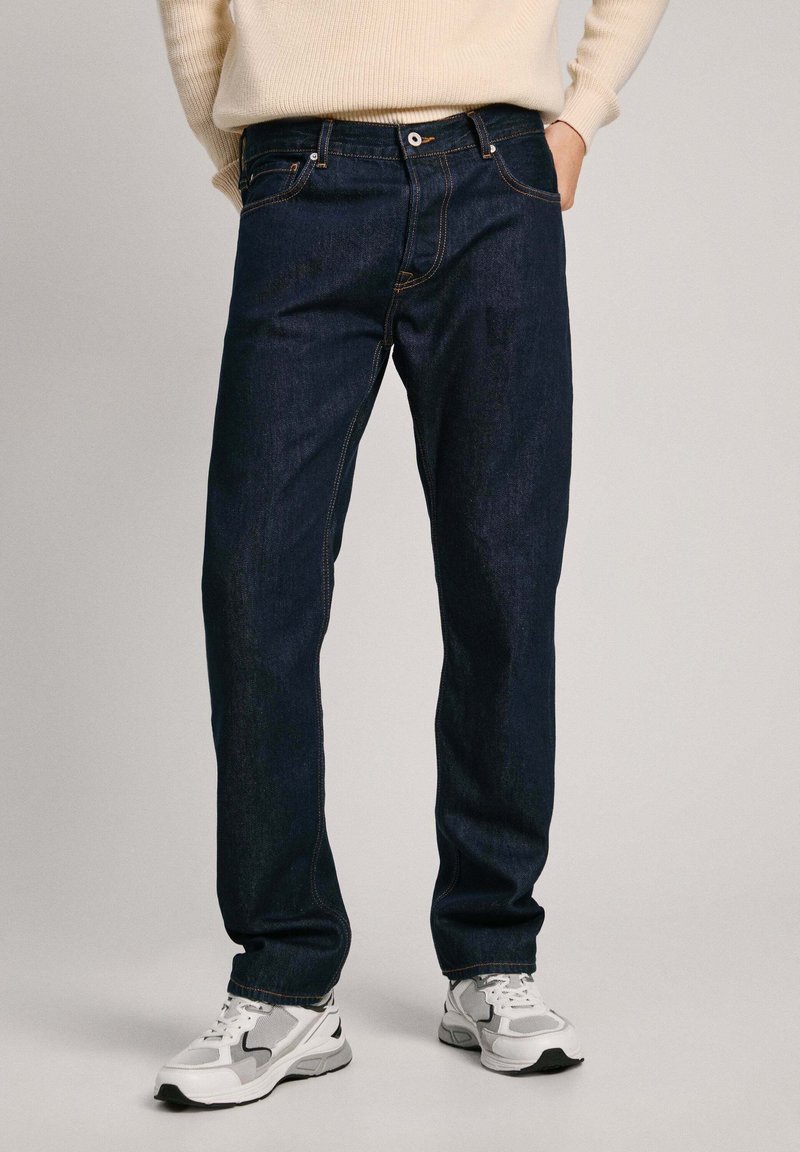 Jeans in denim blu scuro con fit dritto, caratterizzati da cuciture a contrasto, cinque tasche e chiusura con bottone in metallo. Indossati con sneakers bianche.