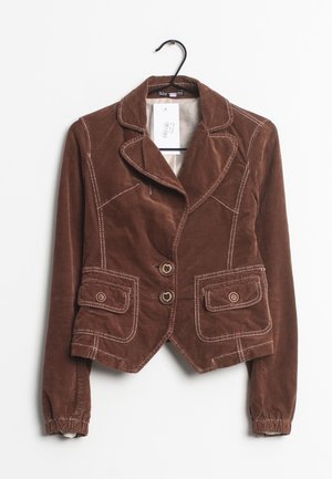 Bruine corduroy blazer met witte stiksels, knopen aan de voorkant, klepzakken en elastische manchetten, hangend aan een zwarte hanger met een wit label.