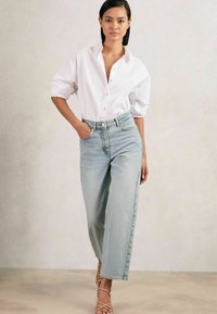 Jeans azzurri a vita alta con una vestibilità morbida, abbinati a una camicia bianca con bottoni. L'outfit presenta maniche arrotolate e sandali beige con lacci.