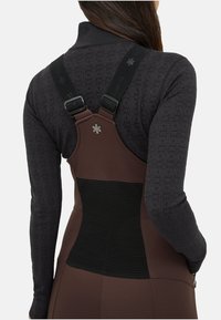 Kvinde iført en brun forklæde-lignende dragt med justerbare sorte stropper over en struktureret mørkegrå langærmet top, vist fra ryggen.