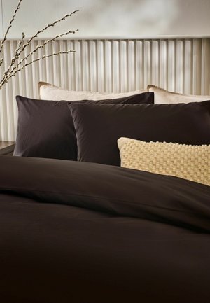 Bett mit dunkelbraunen Laken, beigen Zierkissen, strukturiertem cremefarbenem Kissen und weißem Lattenkopfteil mit Astdekoration an der Seite.