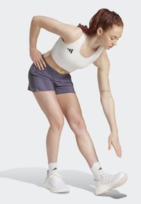 Weißes sportliches Crop-Top mit schwarzen Akzenten, lila Shorts mit seitlichen Schlitzen und weißen Sneakern mit schwarzem Branding. Model führt eine Dehnung durch.