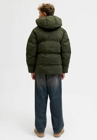 Giacca a vento imbottita verde oliva con colletto alto e cappuccio, caratterizzata da un design trapuntato. Indossata con pantaloni di jeans blu larghi e stivali marrone chiaro.