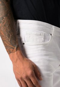 Jeans de mezclilla blanca con un corte ajustado, que cuentan con bolsillos frontales, herrajes de metal y una superficie texturizada. Mano visible, mostrando un tatuaje.