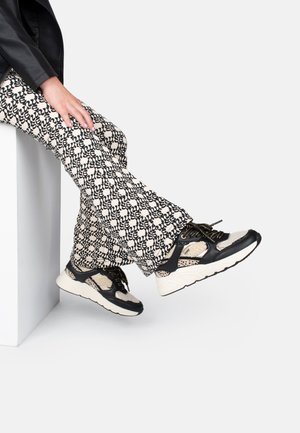 Baskets de sport noires et beiges avec un mesh texturé, des accents en cuir et des lacets dorés, associées à un pantalon à motifs noir et crème.