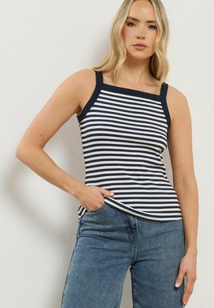 Mujer rubia con top sin mangas de rayas horizontales azul marino y blancas y vaqueros azules, posando con una mano en el bolsillo.