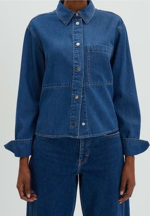 Osoba ubrana w niebieską dżinsową koszulę zapinaną na guziki z przednią kieszenią oraz dopasowane niebieskie dżinsy, stojąca z ramionami rozluźnionymi.