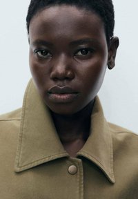 Veste khaki avec un grand col, détail de boutons et tissu texturé, portée par une personne avec des cheveux courts et foncés, sur un fond neutre.