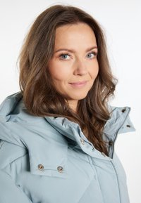 Svijetloplava puffer jakna s visokim ovratnikom, gumbima na pritiskanje i kvadratnim uzorkom. Mekani, teksturirani materijal i detalj sa zatvaračem sprijeda.