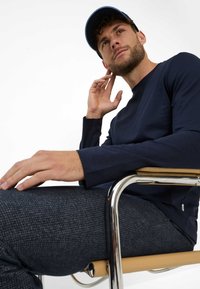 Navyblaues Longsleeve-Oberteil mit Rundhalsausschnitt, kombiniert mit strukturierten, dunkelgrauen Hosen. Die Person sitzt auf einem Stuhl mit Metallrahmen.