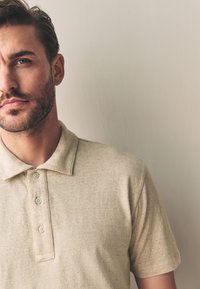 Tricou polo bej din țesătură texturată, având guler și pată cu trei nasturi. Prim-plan al secțiunii superioare a tricoului.